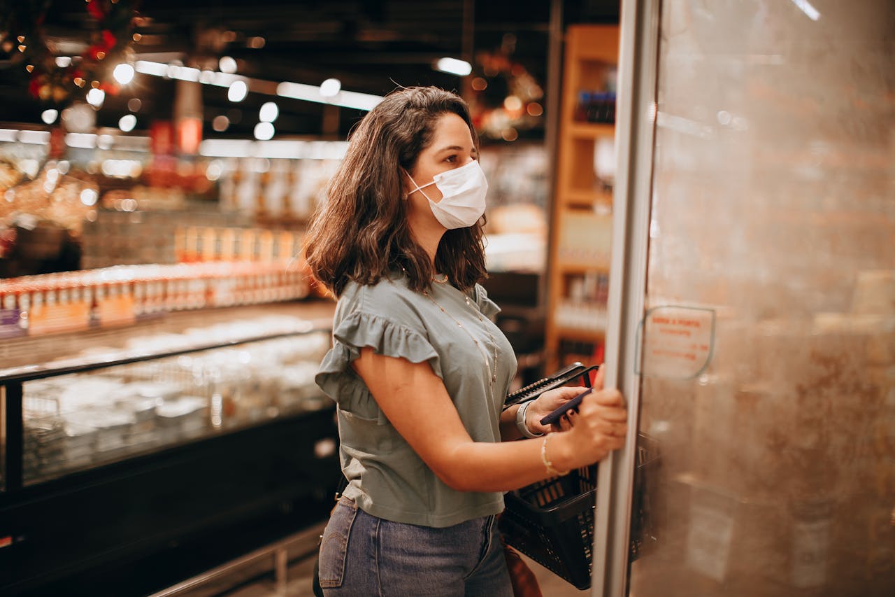 A woman wearing a mask shops in a grocery store, opening a refrigerator.